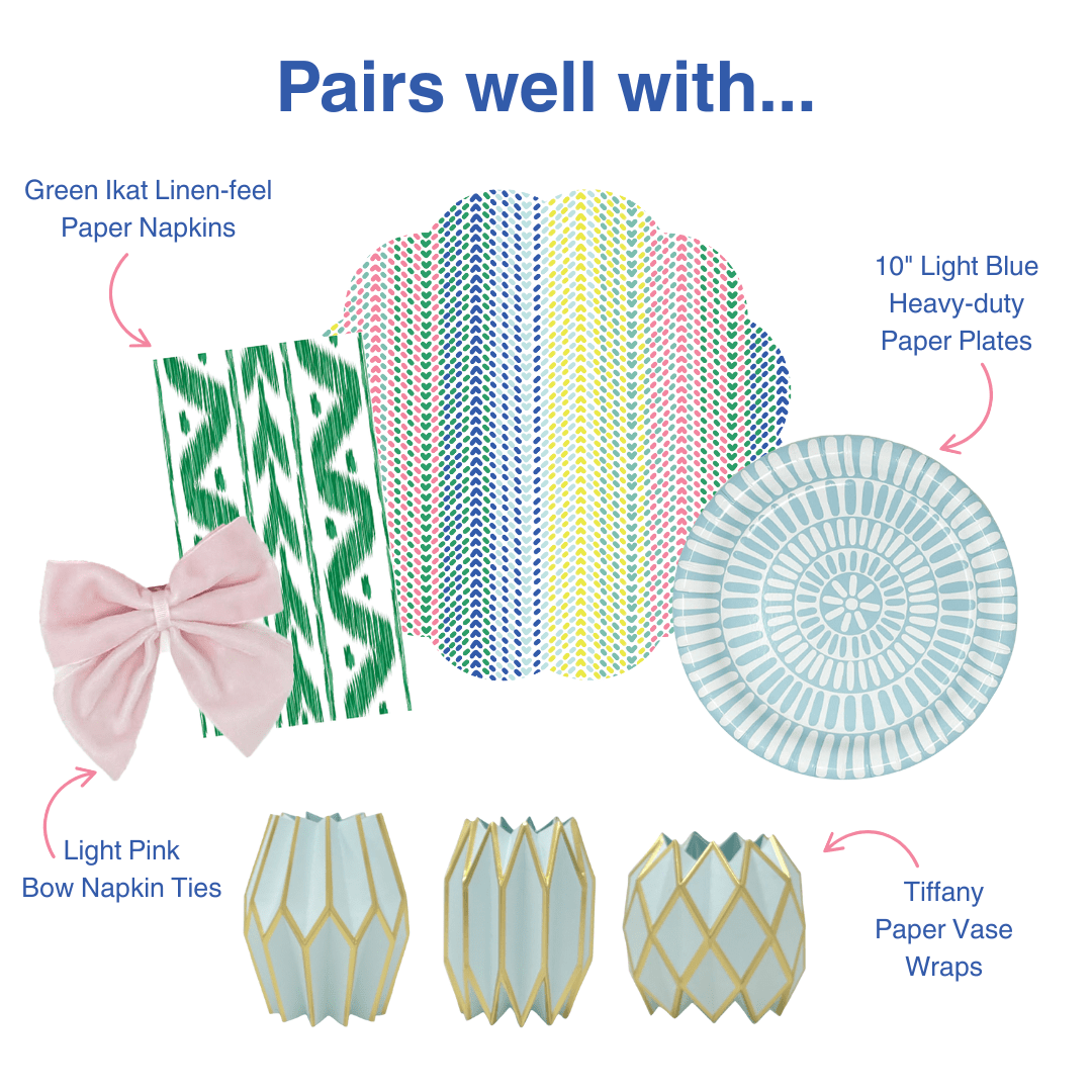 Infographic titled "Pairs well with..." showcasing five complementary Lucy Grymes products: a green ikat linen-feel paper napkin with a blush pink velvet bow napkin tie, a multicolor chevron-dot scallop-edged paper placemat in pink, navy, and yellow stripe pattern, a 10-inch light blue heavy-duty paper plate with a concentric circle dash design, and three Tiffany blue origami-style paper vase wraps with gold geometric detailing. Product names are labeled with pink arrows on a white background.