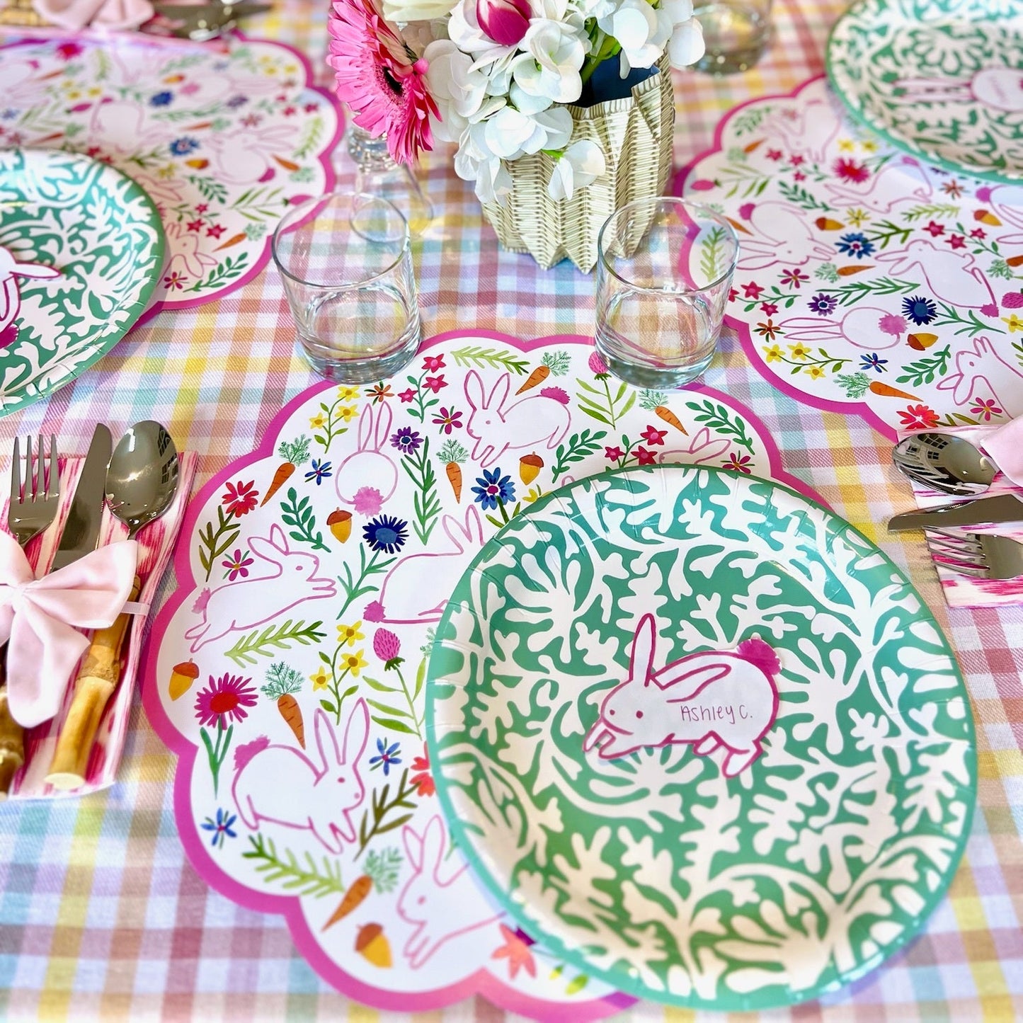 Easter-themed table setting with bunny paper placemats, decorative green plates, napkins, and flowers on a checkered tablecloth.