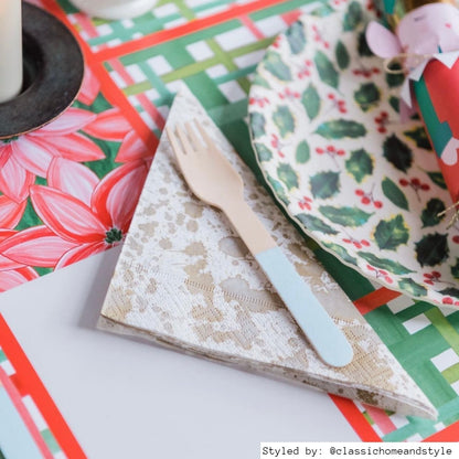Close up of a table setting with red and green plaid paper placemats layered with a red and green holly plate and a cream and white paper napkin folded into a triangle with a wooden fork