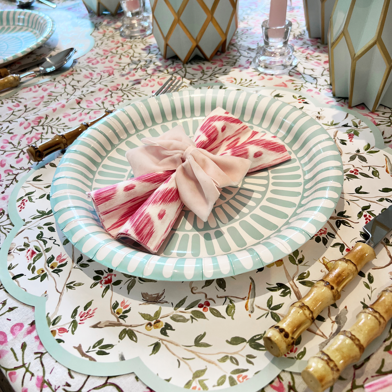 Close-up of a spring place setting with a blue striped paper plate on a scallop-edged charger, topped with a pink ikat napkin tied with a light pink bow, bamboo-handled flatware, and Tiffany paper vase wraps with white florals in the background