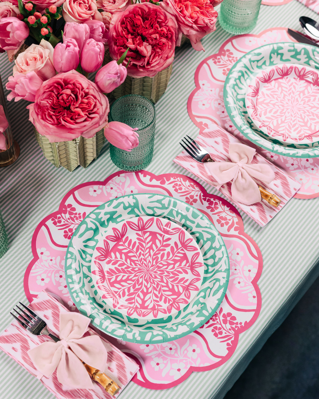 Close-up of a spring tablescape with pink scallop-edged floral placemats, layered teal and pink floral medallion paper plates, pink ikat napkins tied with light pink bows, bamboo-handled flatware, green glassware, and a lush arrangement of coral and blush roses and tulips in a wicker vase.