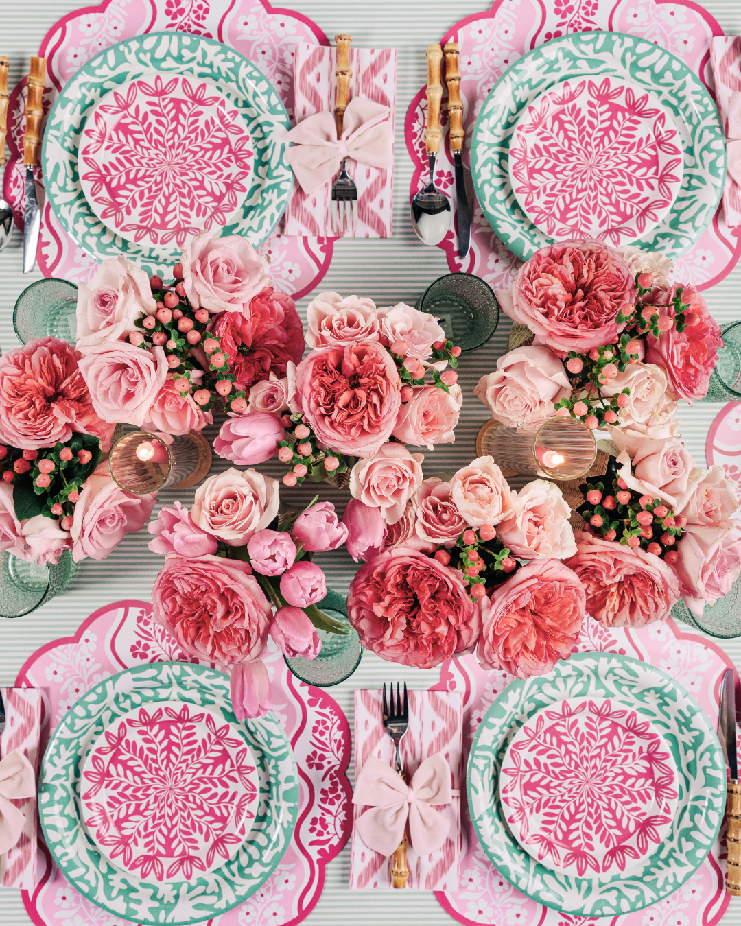 Overhead view of a styled tablescape with six place settings featuring pink scallop-edged floral placemats, layered teal and pink floral medallion paper plates, pink ikat napkins tied with light pink bows, bamboo-handled flatware, and a lush centerpiece of coral and blush roses with green glassware.