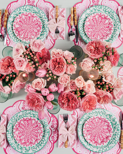 Overhead view of a styled tablescape with six place settings featuring pink scallop-edged floral placemats, layered teal and pink floral medallion paper plates, pink ikat napkins tied with light pink bows, bamboo-handled flatware, and a lush centerpiece of coral and blush roses with green glassware.