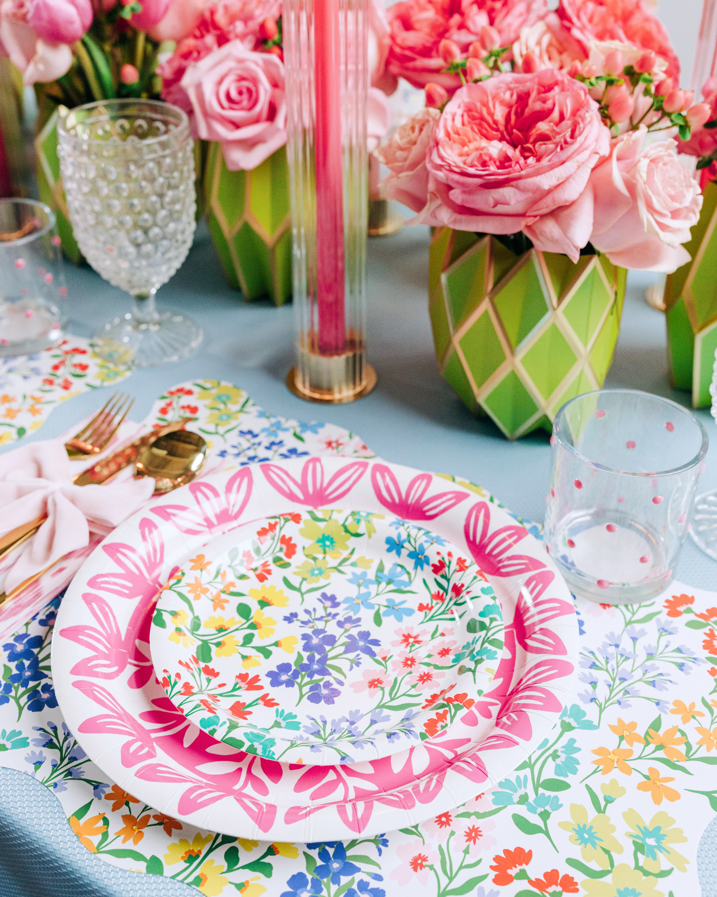 A spring table featuring a scallop-edged wildflower print placemat, layered with a pink floral medallion plate and a smaller Full Bloom wildflower print plate, a pink ikat napkin tied with a light pink bow, gold flatware, and a lush arrangement of coral roses and peonies