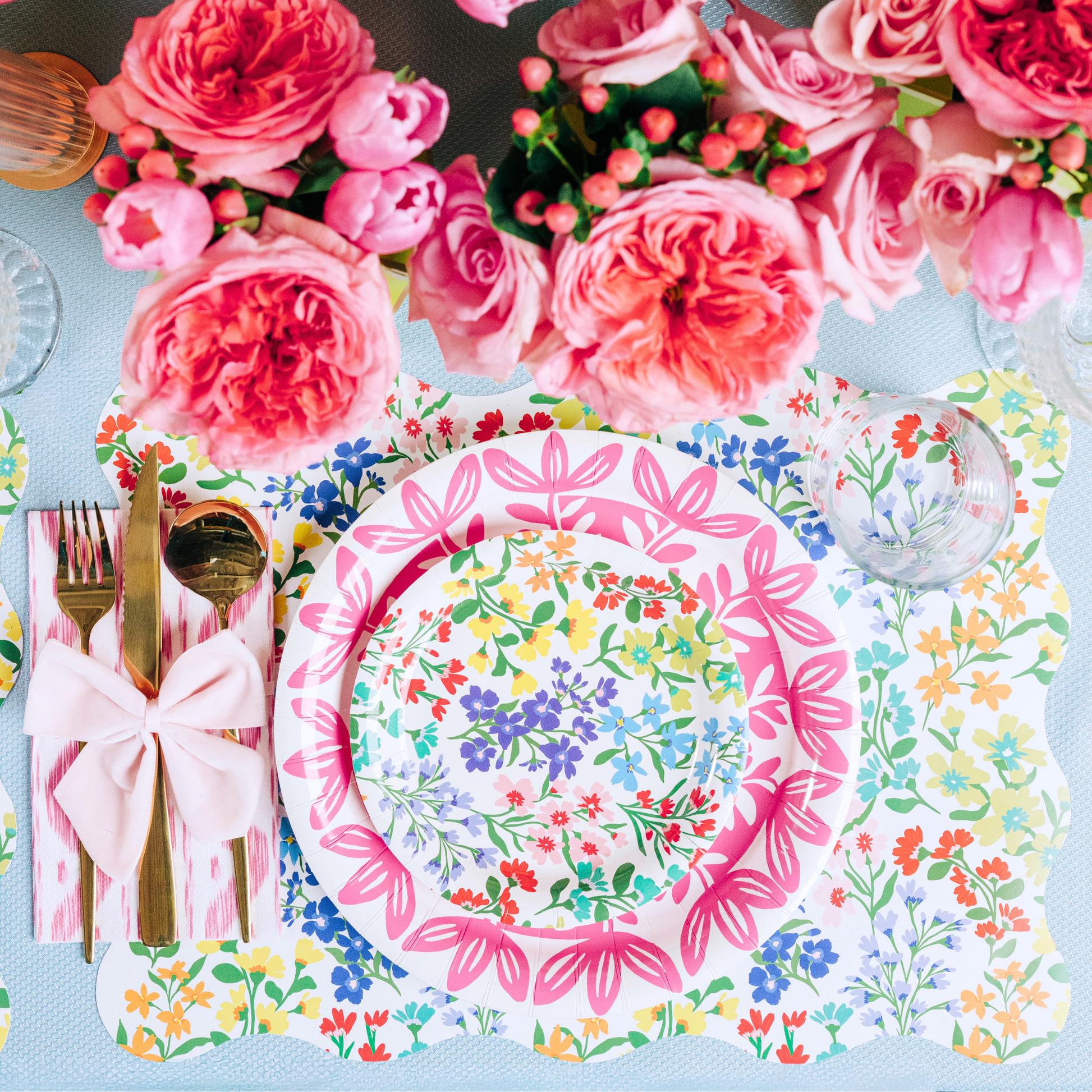 Overhead view of a spring place setting featuring a scallop-edged wildflower print placemat, layered with a pink floral medallion plate and a smaller Full Bloom wildflower print plate, a pink ikat napkin tied with a light pink bow, gold flatware, and a lush arrangement of coral roses and peonies in the background
