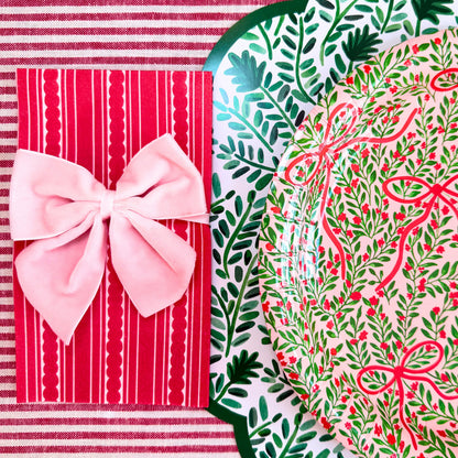 A styled flat-lay of holiday-themed paper goods, featuring a red and white striped paper napkin tied with a pink bow next to a scalloped paper placemat and a paper plate printed with a green wreath and red bow pattern on a red and white striped tablecloth.