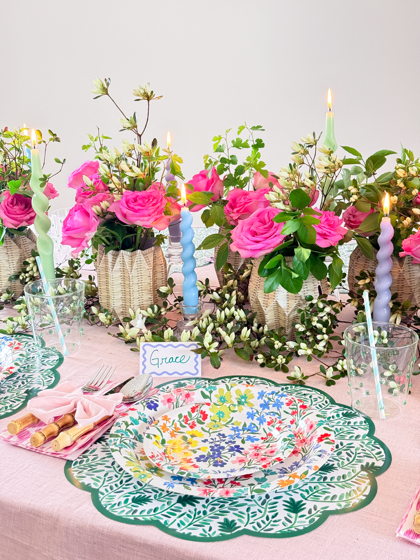 Decorative table setting with floral arrangements, colorful plates, and candles on a light pink tablecloth.