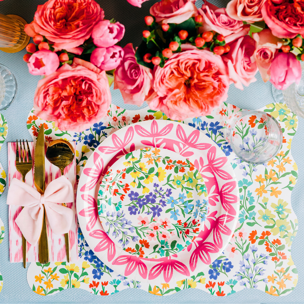Overhead view of a spring place setting featuring a scallop-edged wildflower print placemat, layered with a pink floral medallion plate and a smaller Full Bloom wildflower print plate, a pink ikat napkin tied with a light pink bow, gold flatware, and a lush arrangement of coral roses and peonies in the background