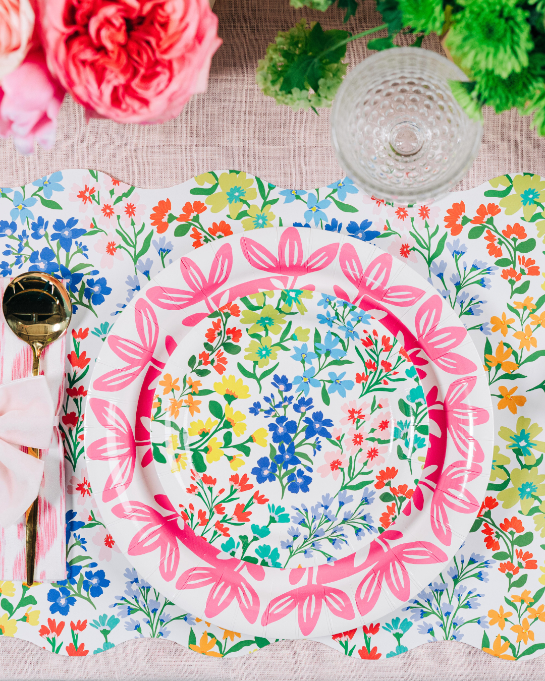 Colorful table setting with a floral scalloped placemat, layered floral and pink-patterned plates, a pink napkin wrapped around gold cutlery, and a glass and flowers above.