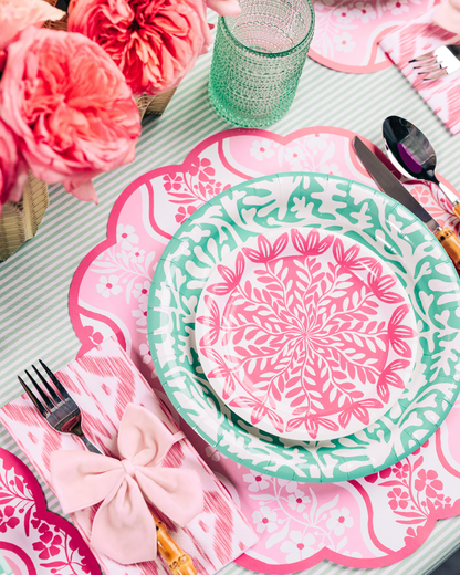 Close-up of a pink and green spring table setting with layered floral-patterned plates on a scalloped placemat, a pink ikat-style napkin tied with a bow, green textured glassware, and pink roses.