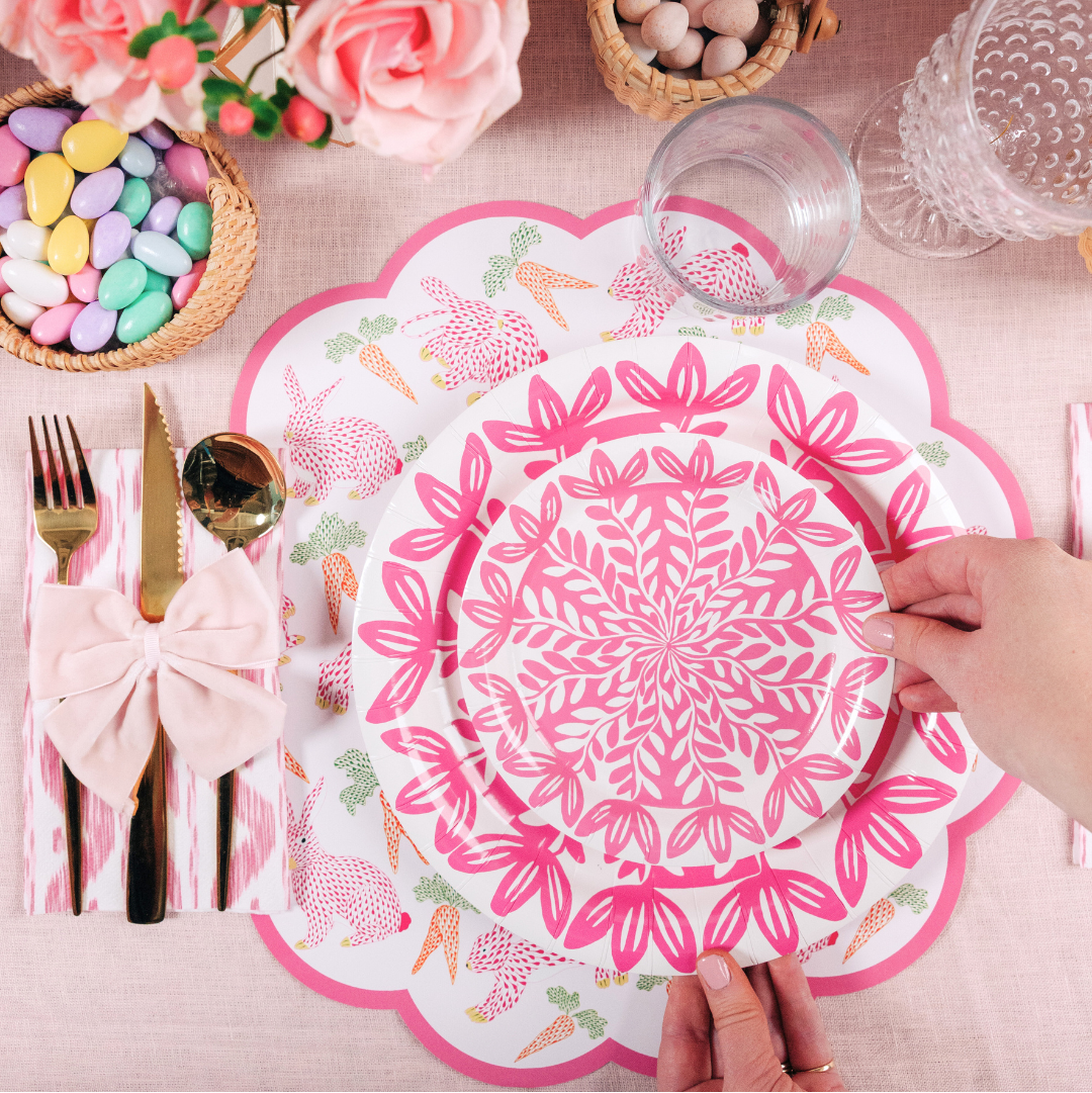 Top-down view of a pink Easter-themed table setting with floral plates, a scalloped placemat with bunnies and carrots, pastel candy in baskets, pink roses, and a hand placing a plate on the table.