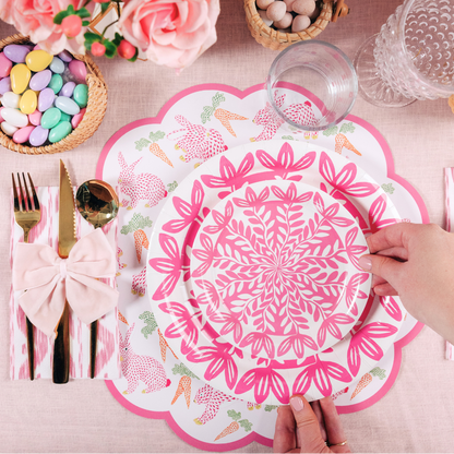 Top-down view of a pink Easter-themed table setting with floral plates, a scalloped placemat with bunnies and carrots, pastel candy in baskets, pink roses, and a hand placing a plate on the table.