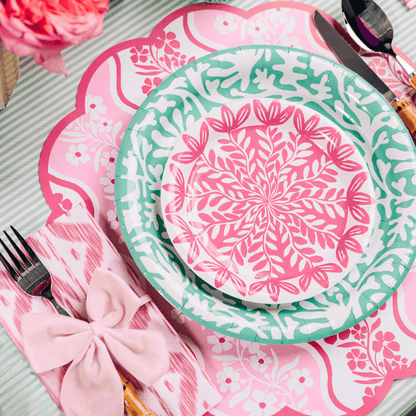 Pink botanical leaf-print dessert plate layered on a teal abstract floral dinner plate, styled on a pink floral scalloped placemat with a pink ikat napkin, pink bow napkin tie, and silver flatware