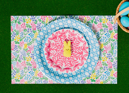 Easter place setting on green grass featuring a colorful wildflower print placemat, layered with a light blue woven cane print plate and a smaller pink floral medallion plate, topped with a yellow Peeps bunny marshmallow, with a basket of blue Easter eggs in the corner