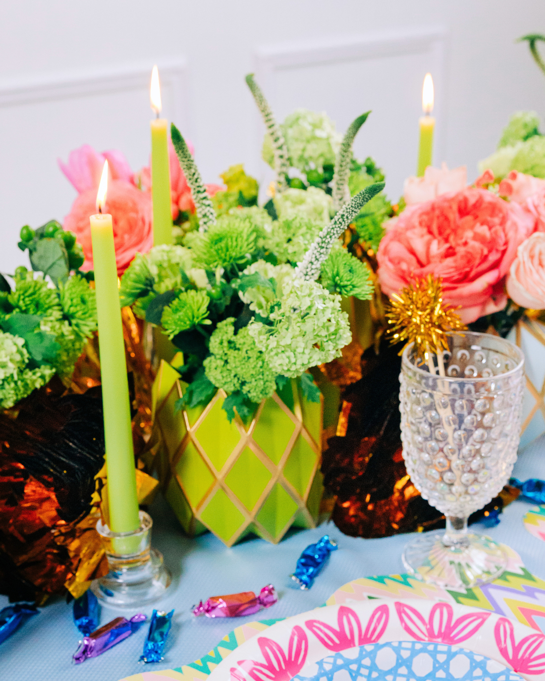 Vibrant party table centerpiece with lime green geometric shaped vase wraps holding pink and coral roses, green hydrangeas, chrysanthemums, tall green taper candles, gold foil accents, and scattered colorful foil-wrapped candies on a blue patterned tablecloth with pink floral plates