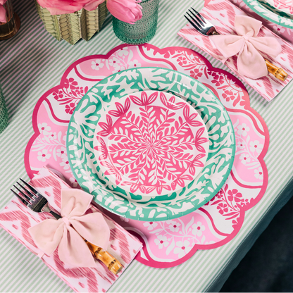 Table setting with floral pink paper placemats, pink and green paper plates, and napkins on a tablecloth.