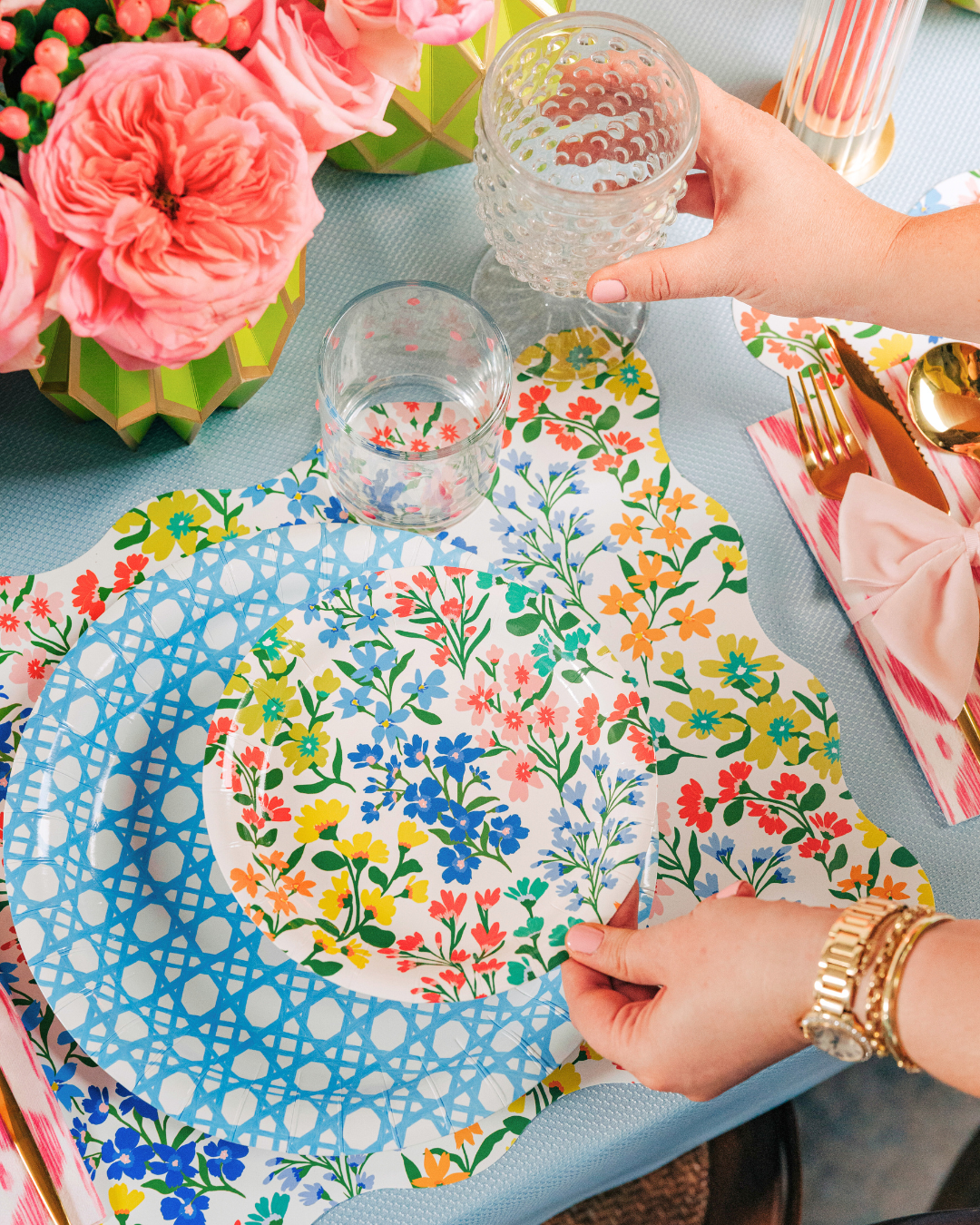 Multicolor wildflower paper plate being placed onto a light blue cane-weave dinner plate, styled on a matching wildflower scalloped placemat with a pink striped napkin and gold flatware