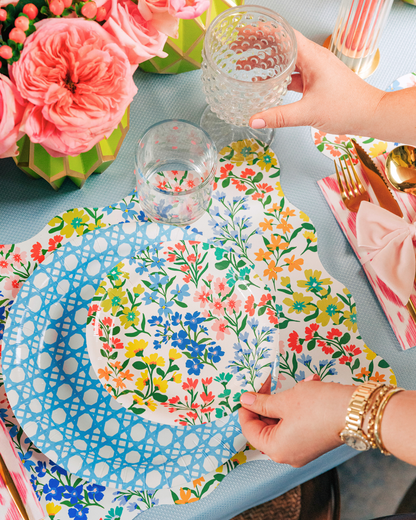 Multicolor wildflower paper plate being placed onto a light blue cane-weave dinner plate, styled on a matching wildflower scalloped placemat with a pink striped napkin and gold flatware