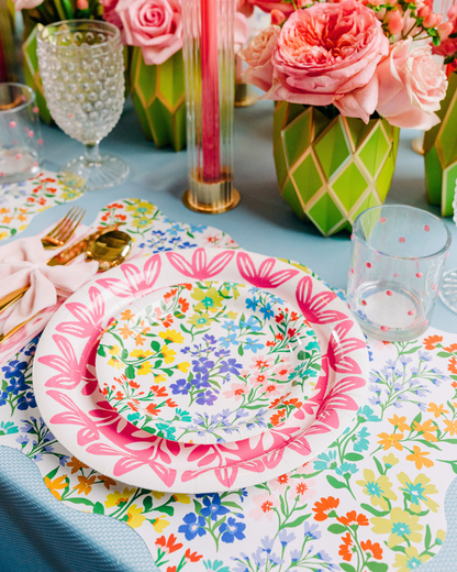 Multicolor wildflower paper plates layered with a pink botanical leaf-print plate, styled on a matching wildflower scalloped rectangle placemat with a pink bow napkin and gold flatware