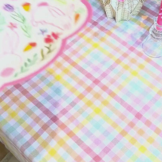 Video showing a pink bunny and wildflower scalloped round placemat being layered with a sage abstract floral dinner plate, styled on a pastel gingham tablecloth