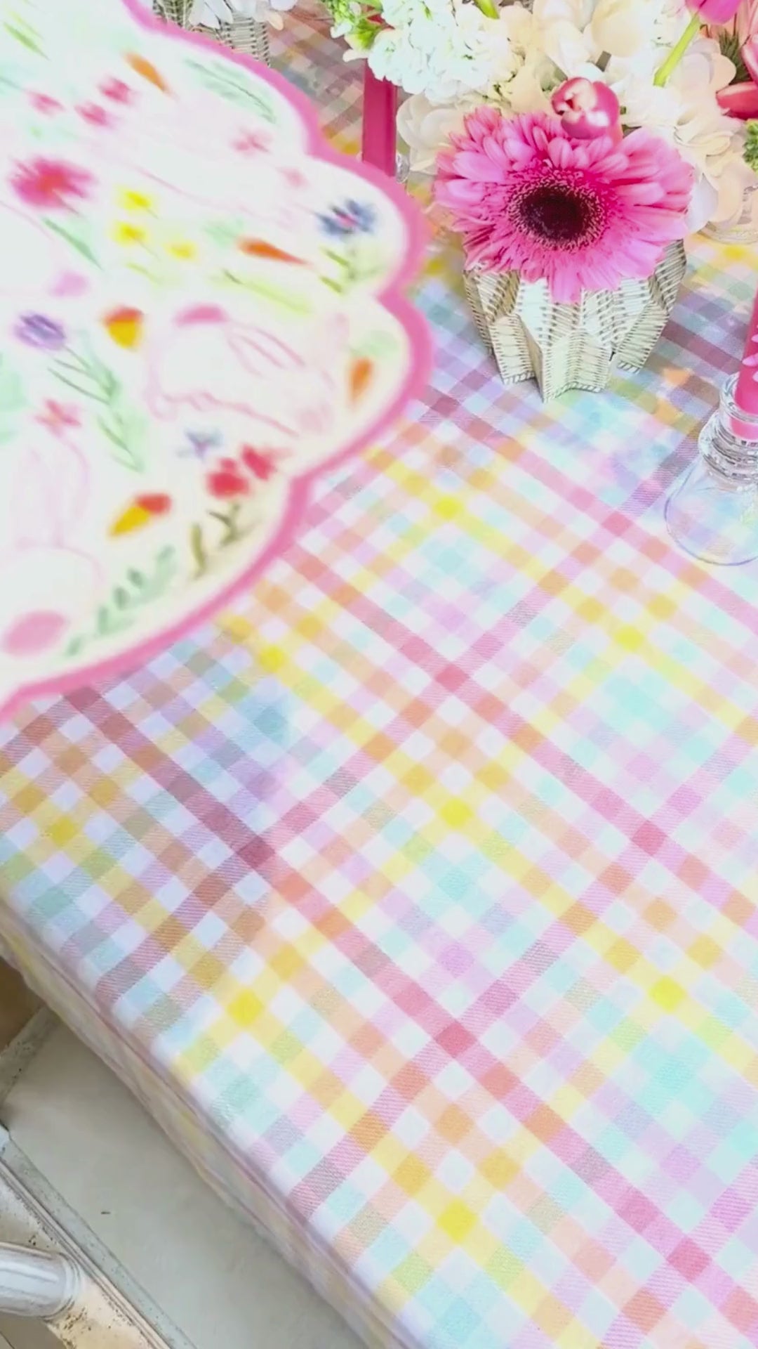 Video showing a pink bunny and wildflower scalloped round placemat being layered with a sage abstract floral dinner plate, styled on a pastel gingham tablecloth