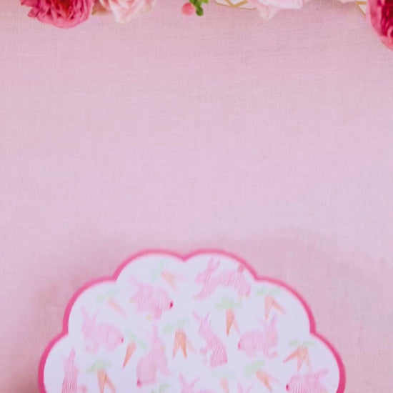 Video showing a round scalloped pink placemat printed with bunnies and carrots being layered with a pink botanical leaf-print dinner plate