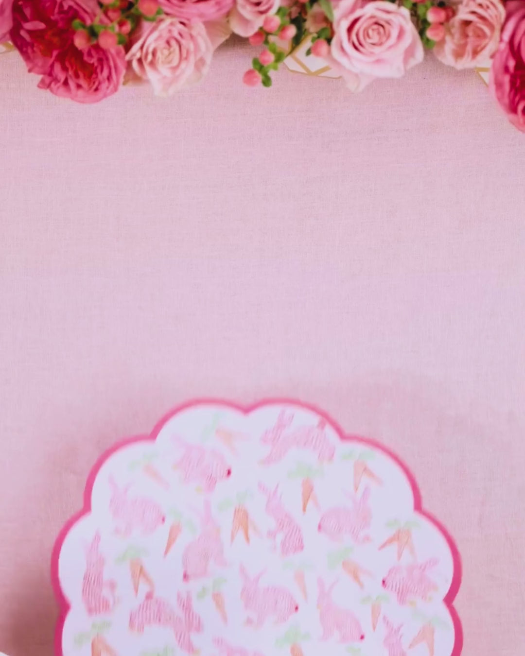 Video showing a round scalloped pink placemat printed with bunnies and carrots being layered with a pink botanical leaf-print dinner plate