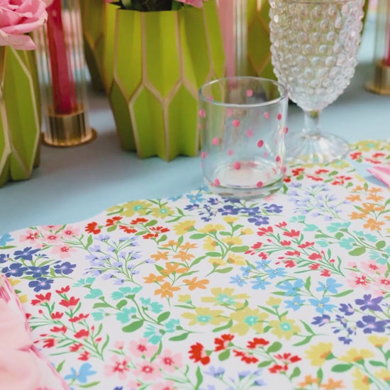A video of a spring table featuring a scallop-edged wildflower print placemat, layered with a pink floral medallion plate and a smaller Full Bloom wildflower print plate, a pink ikat napkin tied with a light pink bow, gold flatware, and a lush arrangement of coral roses and peonies