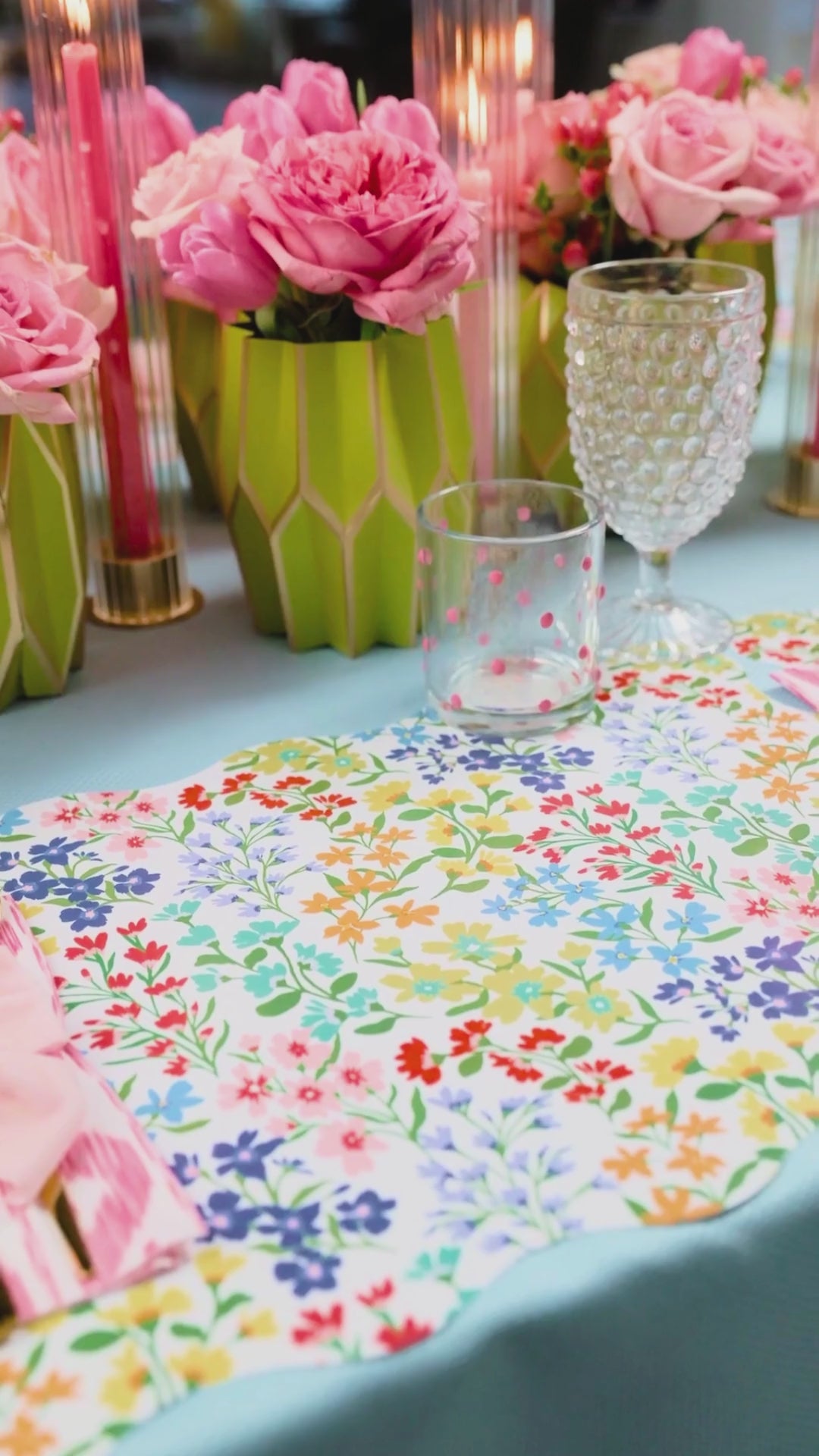 A video of a spring table featuring a scallop-edged wildflower print placemat, layered with a pink floral medallion plate and a smaller Full Bloom wildflower print plate, a pink ikat napkin tied with a light pink bow, gold flatware, and a lush arrangement of coral roses and peonies