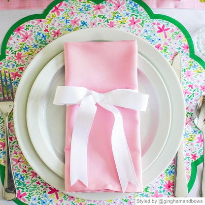 Multicolored floral scalloped round paper placemat layered with white plates and a pink rectangle napkin with a white bow