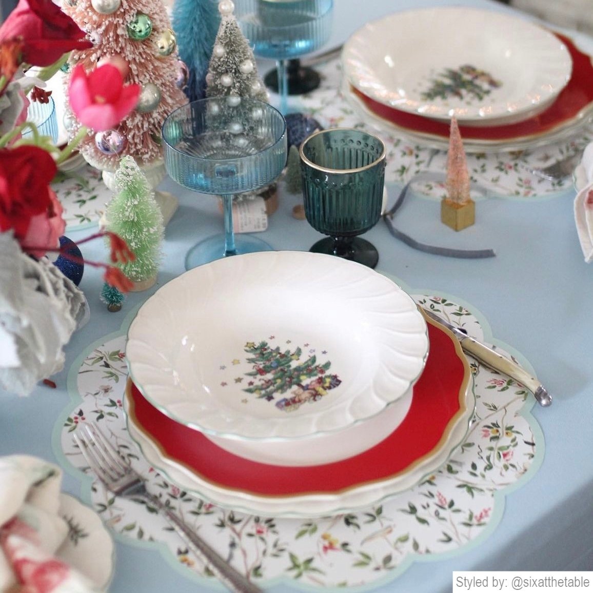Table setting with chinoiserie round scalloped paper placemats layered with red plates and a white plate featuring a Christmas tree on a light blue tablecloth