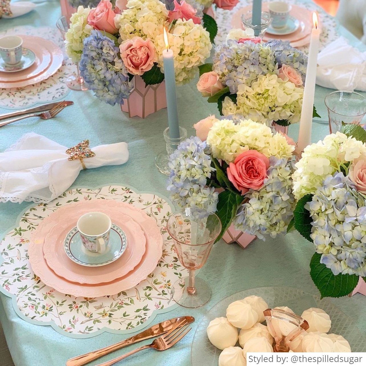 Tea party table setting with white and blue chinoiserie round scalloped paper placemats layered with pink plates and a floral teacup on a blue tablecloth with pink vases and pink, red and blue spring flowers