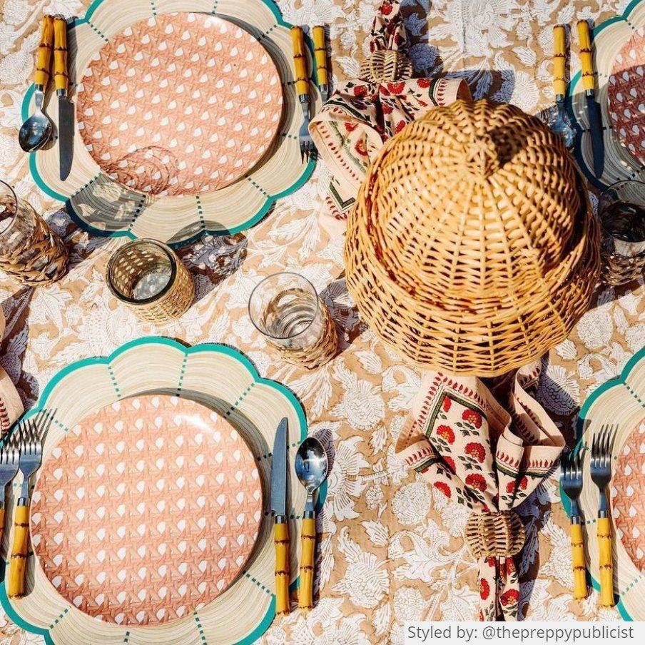 Table setting with round scalloped wicker paper placemats layered with coral fretwork dishes on a brown floral tablecloth with various rattan table decor pieces