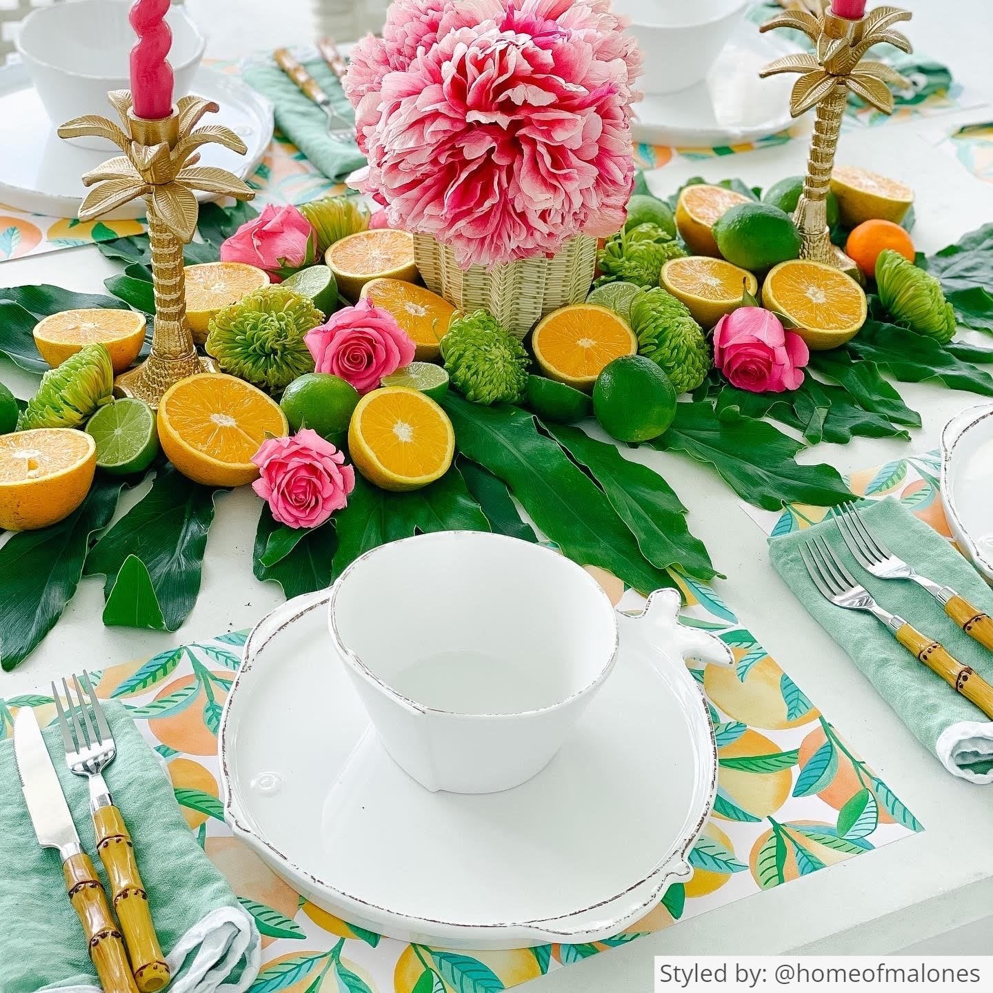 Table setting with paper placemats featuring oranges layered with white dishes and tea cups cut open oranges and green leaves as a centerpiece along with a wicker patterned paper vase wrap with pink flowers