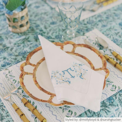 White floral paper placemats layered with a bamboo plate and silverware and a monogrammed napkin on a blue and green floral tablecloth