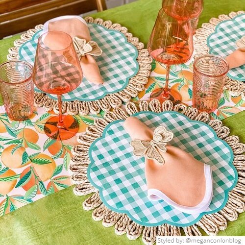 Table setting with rattan chargers and gingham paper placemats with an orange patterned paper tablerunner on top of a bright green tablecloth
