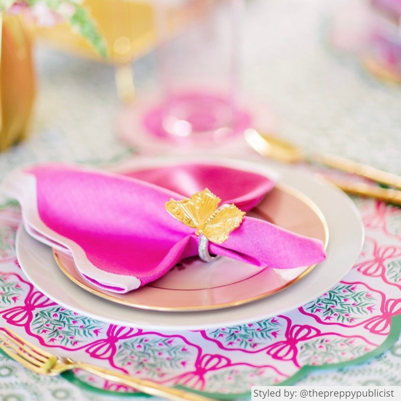 Place setting with pink and green scalloped round paper placemats layered with white and gold plates and a pink napkin tied with a gold butterfly