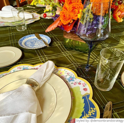 Round scalloped paper placemat featuring a tobacco leaf pattern layered with a white plate and white napkin on a dark green tablecloth