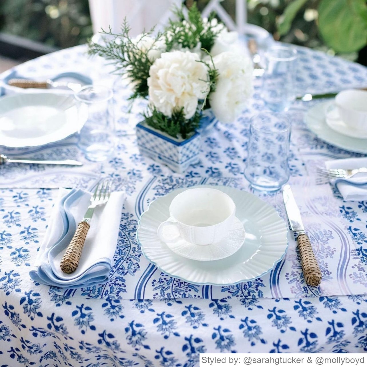 Table setting with blue and white holly paper placemats layered with white dishes and a teacup on a blue and white floral tablecloth with a white floral centerpiece