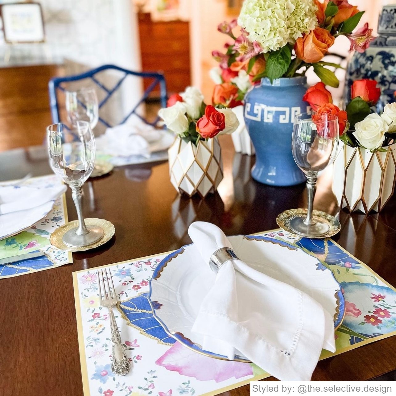 Place setting with a tobacco leaf paper placemat layered with a tobacco leaf plate and white napkin with a silver napkin ring on a wooden table with white and gold vases filled with orange and white flowers