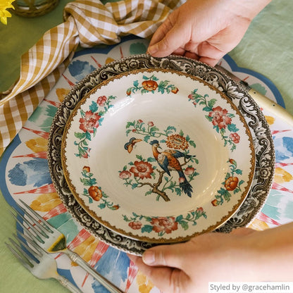 Multicolored feather scalloped paper placemat with a brown gingham napkin on a light green tablecloth with someone placing a stack of plates on top