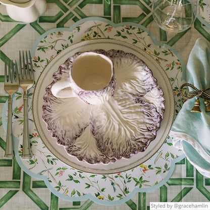 Place setting with a round scalloped chinoiserie paper placemat layered with a purple cabbage plate and teacup on a green fretwork tablecloth