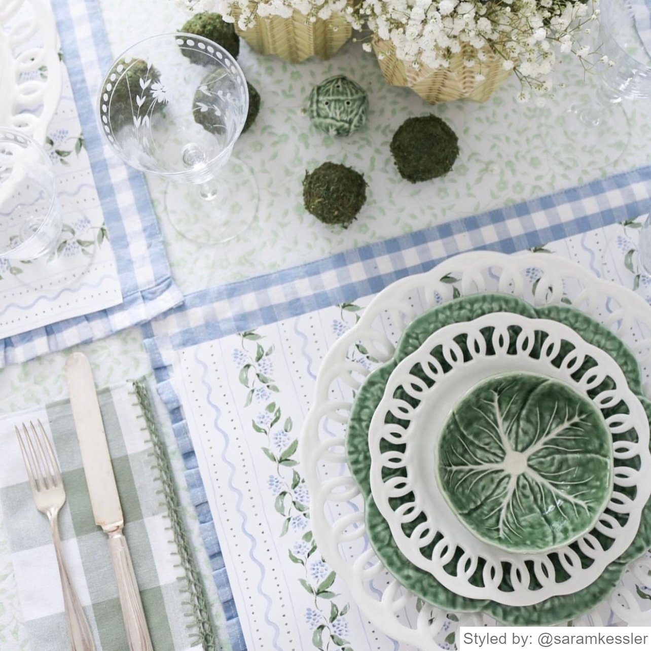Place setting with white floral paper placemats layered with white and green dishes on a blue gingham placemat and green and white tablecloth