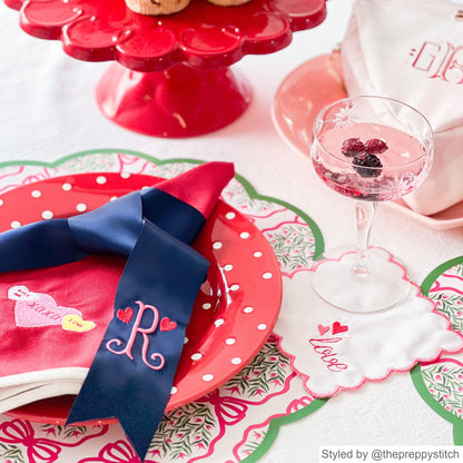 Place setting with pink and green scalloped round paper placemats on a light pink tablecloth layered with a red plate with red polka dots and a pink napkin with a monogrammed blue ribbon napkin ring