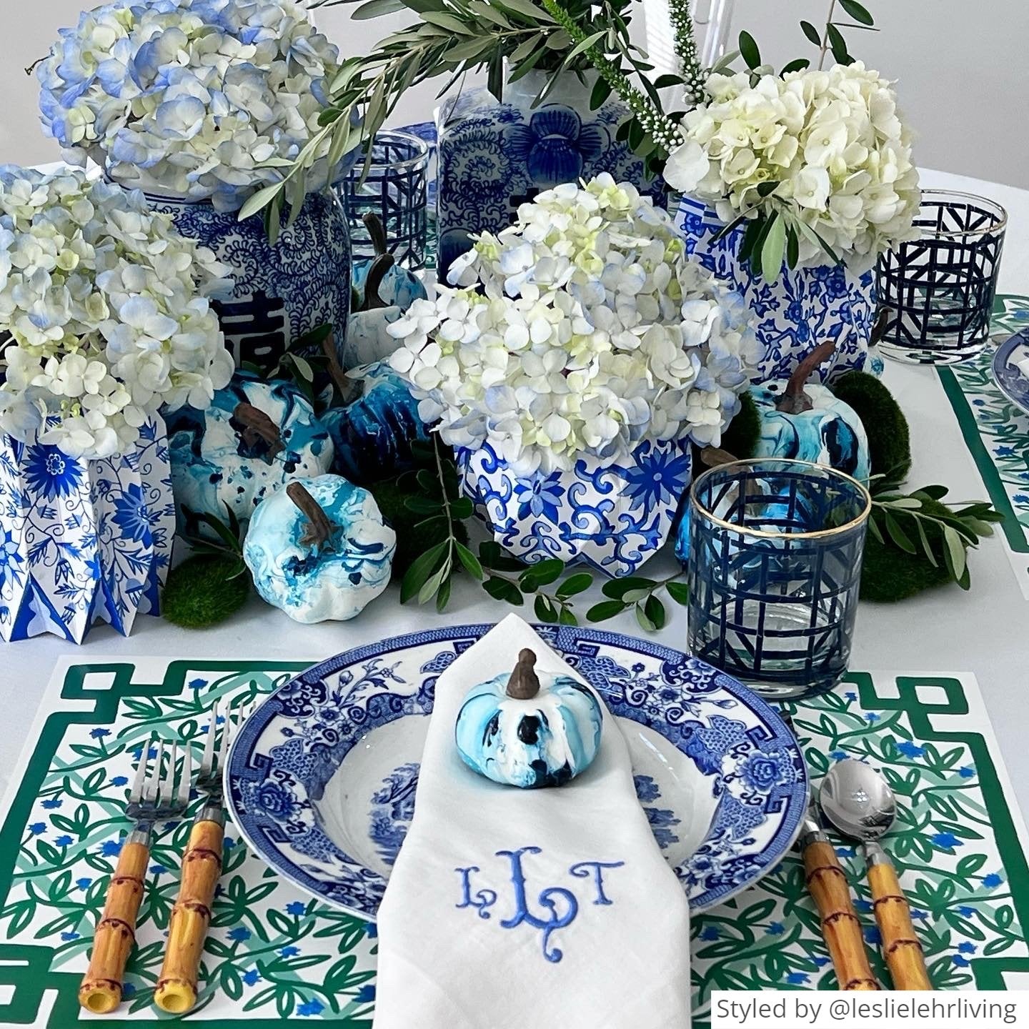 Place setting with green and white paper placemat layered with a blue and white plate, white napkin and blue mini pumpkin on a white table with blue and white vases and flowers