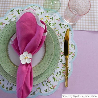 Place setting with a chinoiserie round scalloped paper placemat on a pink tablecloth layered with green and white dishes and a pink napkin with a flower napkin ring