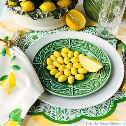 Round scalloped vine paper placemat layered with green and white dishes and yellow jelly beans and lemons on a yellow gingham tablecloth