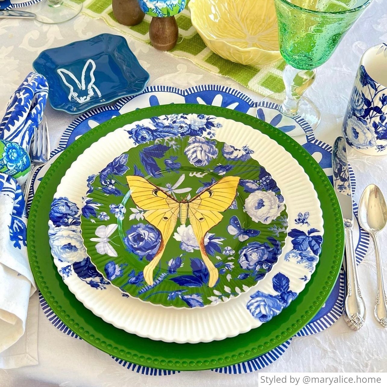 Place setting with a blue and white scalloped round paper placemat layered with green and white dishes with a yellow butterfly