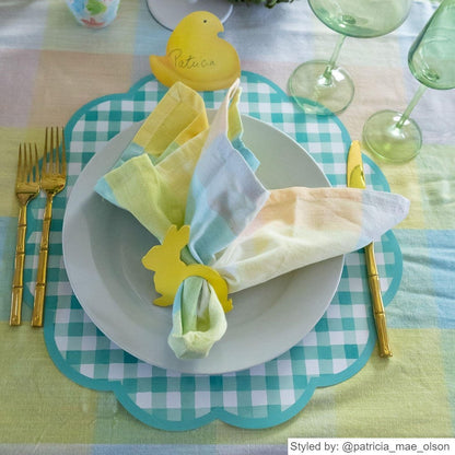 Easter place setting with a green gingham scalloped round paper placemat layered with a white plate and a tie dye pastel napkin tied through a yellow bunny napkin ring