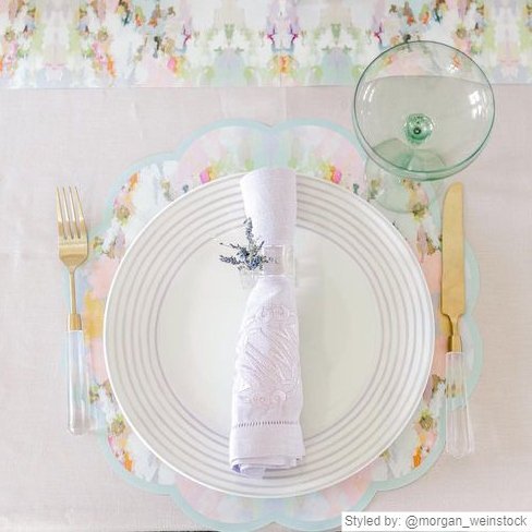 Place setting with blue and pink watercolor scalloped paper placemat layered with a white plate and white napkin on a light pink tablecloth